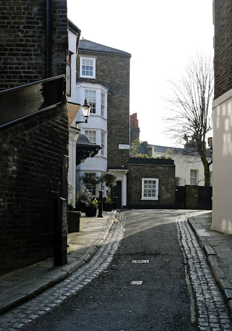 A narrow residential street in West Hampstead showing cobblestone paving alongside smooth asphalt roadways, lined with brick and stucco buildings featuring large windows and exterior lighting fixtures