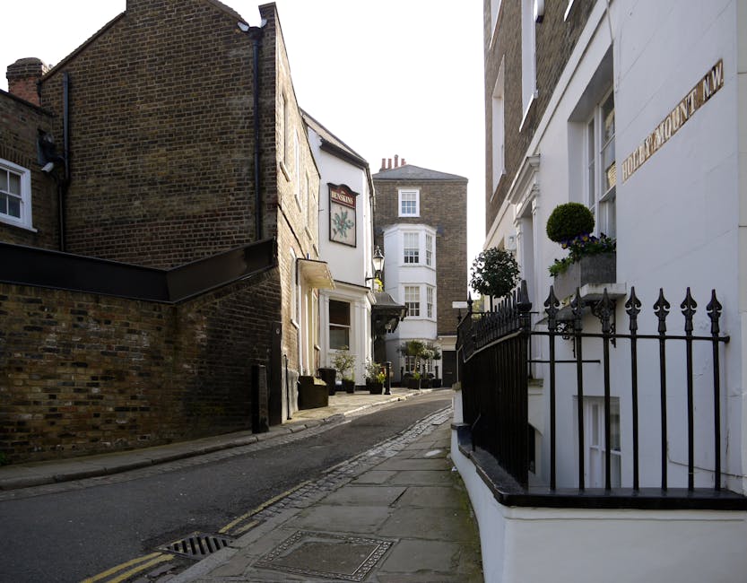 A narrow residential street in West Hampstead showing cobblestone paving alongside smooth asphalt roadways, lined with brick and stucco buildings featuring large windows and exterior lighting fixtures
