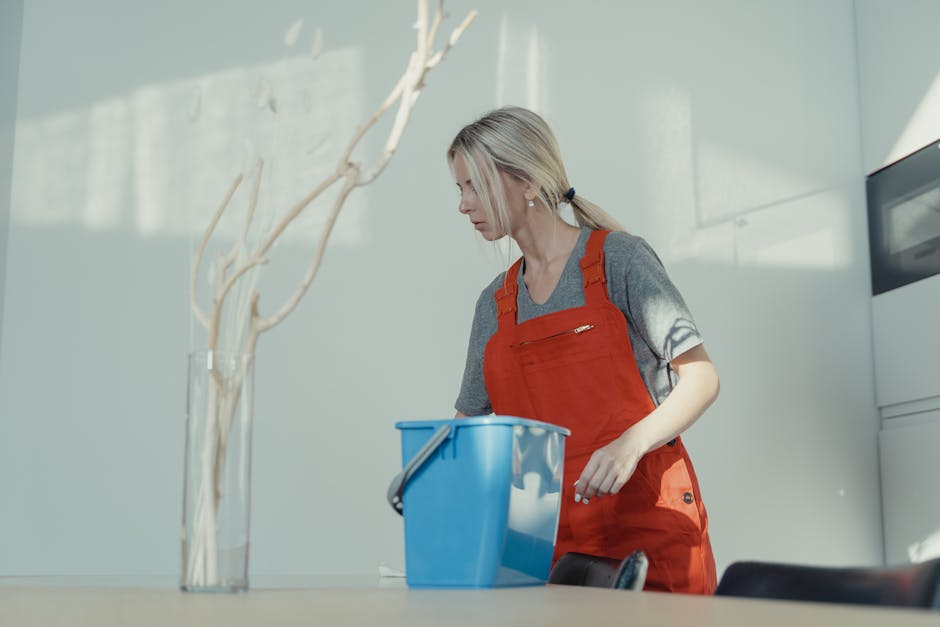 A professional cleaner from Cleaners West Hampstead engaged in domestic cleaning, holding a plastic bucket filled with cleaning tools such as sponges, spray bottles, and cloths. The cleaner, dressed i