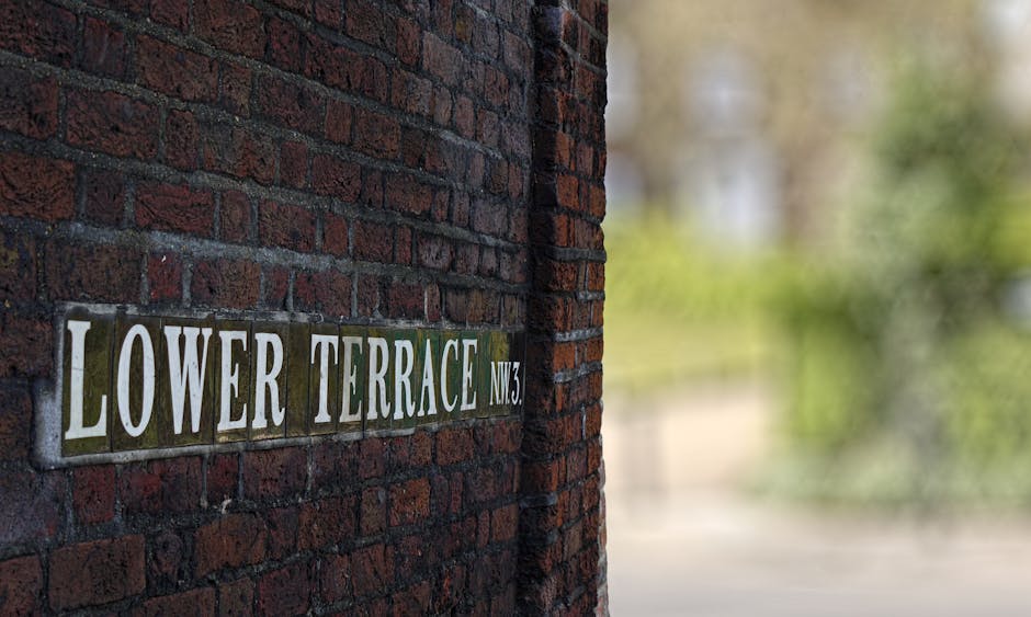 Close-up of a street sign mounted on a red brick wall, reading 'LOWER TERRACE NW3' with white lettering on a dark green background. The wall shows a textured surface composed of weathered bricks with 